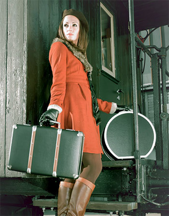nostalgiekoffer Eine Frau in einem orangefarbenen Mantel und mit einem Koffer in der Hand steht auf den Stufen eines Oldtimer-Wagens mit einer runden Hutschachtel.