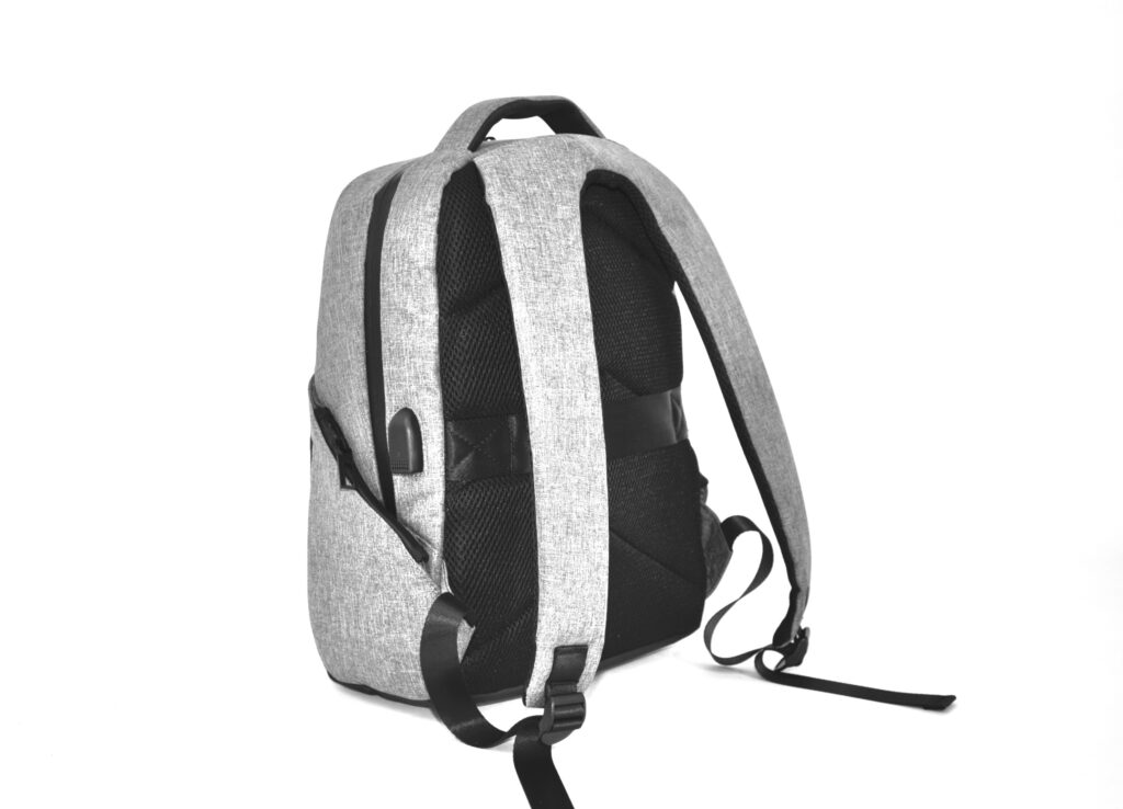 A light gray backpack with black straps and a handle, shown from the back and side on a white background. Inspired by aluminum boxes, this backpack has a visible USB charging port and allows for customization.