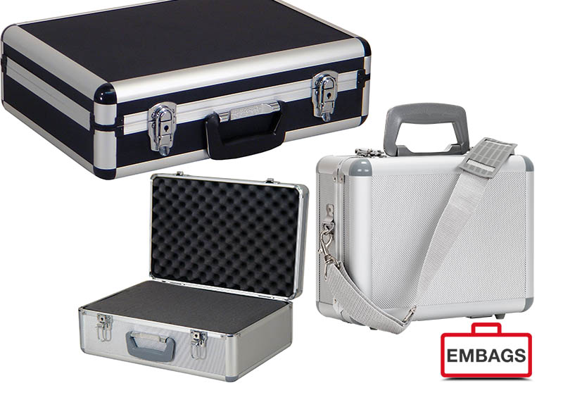 Embags Drei robuste, silber-schwarze Aluminiumkoffer mit Metallkanten; einer ist offen mit Schaumstoffpolsterung für individuelle Anpassungen, ein anderer ist geschlossen mit Griff und Schulterriemen. Rechts unten erscheint das EMBAGS-Logo mit einem roten Koffersymbol.