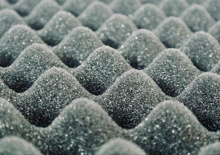 Primer plano de espuma de cartón de huevos gris oscuro con una superficie ondulada y texturizada. Los picos y valles de la espuma forman un patrón repetitivo y dan impresión de profundidad. El material parece blando y poroso, y suele utilizarse para aislamiento acústico o tapicería.