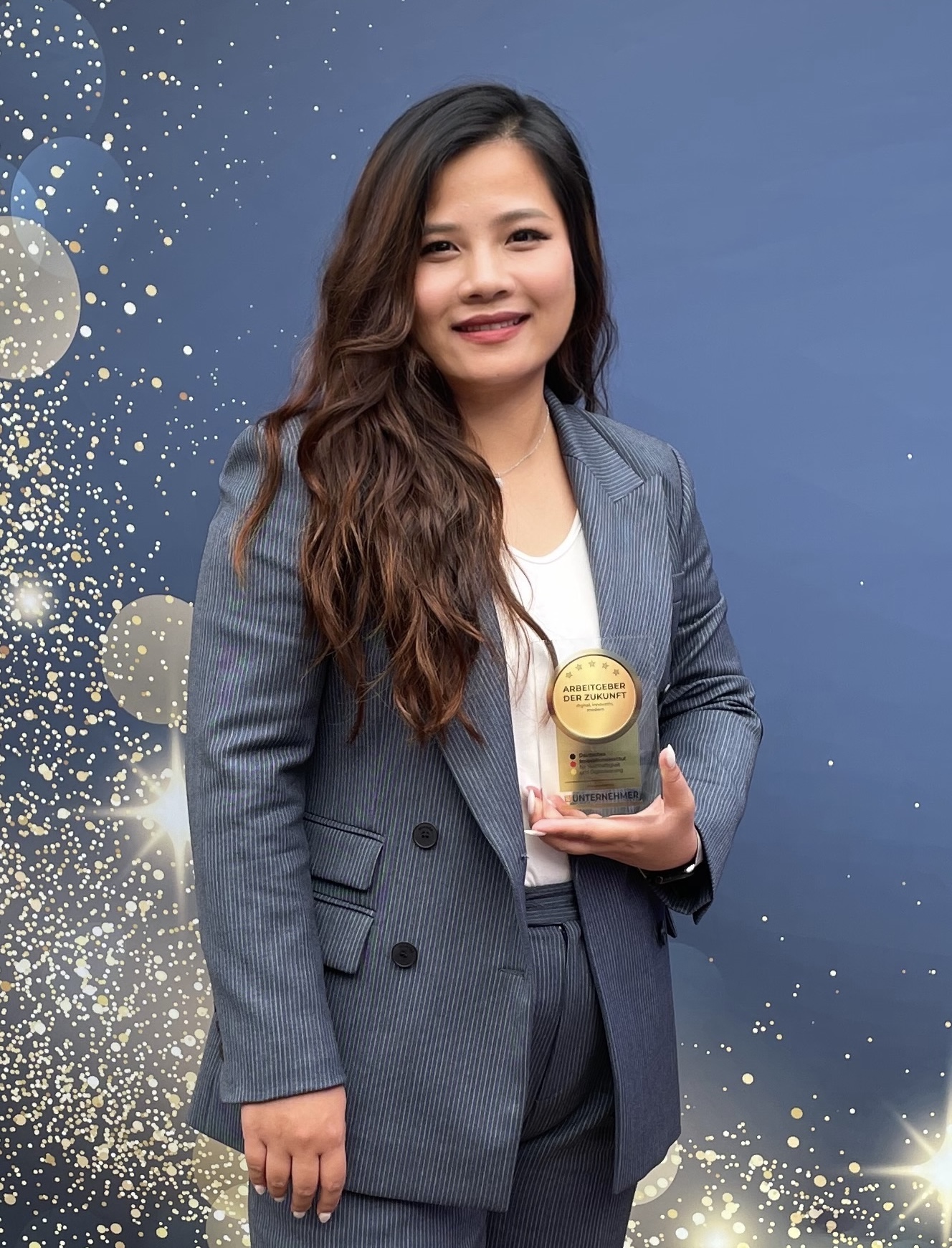 Eine Frau im Kostüm hält eine goldene Trophäe in der Hand und lächelt in die Kamera, während sie vor einem blauen Hintergrund mit Glitzer steht.